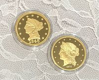 Two gold tone replica coins encased in clear plastic placed on lace fabric, showing obverse sides with Liberty portraits and dateyears.
