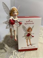 Front view of the Hallmark Keepsake Very Merry Christmas Fairy hanging ornament with the original box behind it, showing the fairy in a red and white outfit with wings and holding a wand.