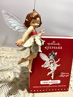 Front view of the fairy ornament suspended showing red bodice, white petal skirt, translucent wings, and original red box in background