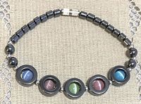 Close-up view of the hematite bead bracelet lying flat on a textured surface, showing the arrangement of cat's eye circular accents and cylindrical hematite beads with metallic clasp.