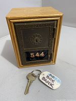 Front and angled view of the wooden piggy bank showing metal door with box number 544 and included key with tag