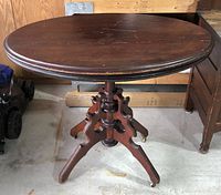 Front and side view of the oval antique parlour table showing carved pedestal base with four legs on casters and aged wood surface.