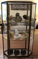 Front and side view of the brass and glass display case with wooden table and living room background. Shows all 9 Wade figurines inside across three shelves.
