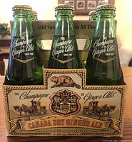 Photo of original cardboard carrier holding six green glass bottles, with vintage-style advertising graphics on carrier.