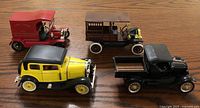 Four collectible Ford Model T diecast cars displayed together on a wooden surface: a red fire chief truck, a wagon with wood paneling, a black flatbed truck, and a yellow and black sedan.