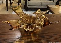 Golden amber art glass display dish on wooden table, front view showing flowing organic shape and glossy finish.