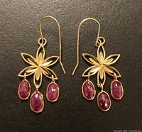 Earrings on black background showing flower-shaped 14K yellow gold design with three oval ruby drops each