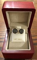 Pair of round black diamond stud earrings set in 14K yellow gold displayed in a red wooden jewelry box with velour interior.