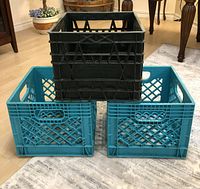 Three plastic milk crates arranged: one black crate stacked on top of two aqua blue crates placed side by side on floor.