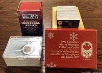 Three complete sports card sets in original packaging displayed on wood surface: NHL 75th Anniversary in clear plastic case with visible white and silver card, World League Football Inaugural Series maroon box, 1992 Canadian Winter Olympic cards in red box with Canadian maple leaf and Olympic rings.