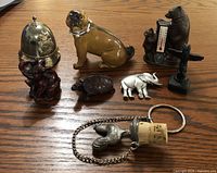 Full view of all eight small collectibles featuring porcelain dog, metal bobbies helmet, tortoise, elephants, bear thermometer, and chain item as arranged on wooden surface.