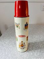 Off-white vintage Thermos vacuum bottle standing upright with red plastic lid and colorful provincial crests including Canadian coat of arms.