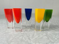 Front view of five colored shot glasses in red, orange-red, blue, yellow, and green, showing their tall square shape with clear glass bases.