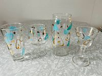 Four Federal Glass Atomic Amoeba pattern glassware pieces displayed: brandy snifter, stemmed cocktail glass, hi-ball glass, and jar/vase. All decorated in turquoise and gold mid-century abstract amoeba shapes.