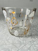 Clear glass ice bucket showing white and gold Atomic Amoeba pattern scattered over the exterior surface, displayed on a textured light surface.