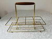 Photo showing gold-tone metal beverage caddy with wooden handle and grid design for glass placement, on a light patterned surface.