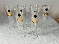 Set of six pilsner glasses decorated with bowling pin and bowling ball graphics in black and gold, with gold rims, standing on a gray patterned surface.