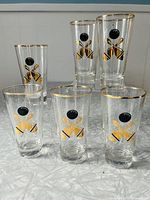 Photo showing six clear glass tumbler glasses arranged in two rows, displaying black and gold bowling pin and ball graphics with gold rims.