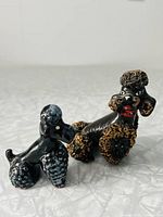 Pair of ceramic poodle figurines side by side on a light surface, showing different sizes and textured curly fur detail.