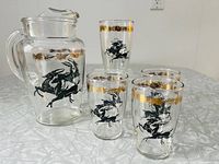 Full set showing one clear glass pitcher and four matching clear glasses decorated with black leaping gazelles and gold accent bands on rims, positioned on a reflective surface.