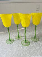 Set of four yellow flower-themed artisan hand-blown wine glasses with green twisted stems and bases on a gray patterned surface.