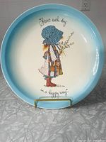 Full view of the vintage Holly Hobbie plate displayed on a stand showing the pale blue gradient border, central character illustration, and text.