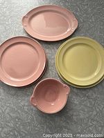 Collection of Lu-Ray Pastels dishes including pink platter, small pink bowl, yellow and pink plates arranged on a silver-gray textured surface.