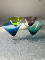 Four jewel-toned martini glasses placed on a surface with natural light highlighting their colors and shapes.