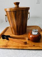 Wood ice bucket with lid, wood tongs, wooden bar tray, and round table lighter arranged on bar tray