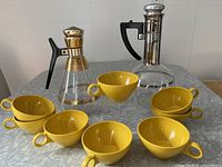 Photo showing two vintage Inland Glass coffee carafes, one with gold bands and one with silver bands, alongside eight yellow plastic cups arranged on a table.
