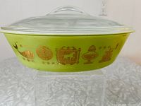 Front and side view of oval green Glasbake serving dish decorated with gold metallic Americana-themed pattern with clear glass lid on top.