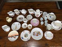 Overview of entire lot of antique child’s and doll china pieces displayed on a wooden table showing cups, plates, creamers, teapots, and small figurines.