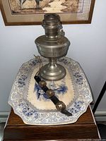 Full view of metal Aladdin lamp sitting on blue and white platter along with leather strap with bells on wooden surface.