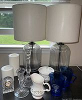Photo showing two glass-base lamps with white fabric shades, two white battery-operated pillar candles, marked Tiffany crystal candlestick, two Irish China pieces with green shamrocks, two vintage blue Shirley Temple creamers, and part of a basket.