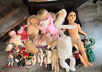 Photo shows an assortment of about 20 dolls including a large nude doll with dark curly hair, a Cabbage Patch Kid doll, various smaller dolls in vintage and traditional clothing, and an antique doll trunk partly visible.