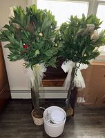 Two faux greenery topiary trees with small lights, wrapped at the base with white tulle, standing on floor next to four planters, different styles and sizes, and a pair of gloves visible
