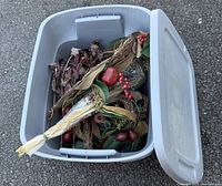 Open gray plastic tote filled with decorative garland featuring artificial greenery, red berries, pinecones, and faux fruit accents.