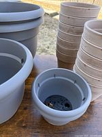 Several gray plastic planters stacked, featuring built-in drainage holes inside. Beige terra cotta planters stacked next to them, showing smooth matte finish and some soil residue inside indicating light use.
