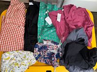 Various women's clothing items displayed flat on a yellow surface including colorful blouses, shirts, pants, and a dress.