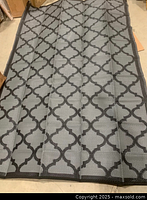 Photo showing the full surface of the outdoor rug laid out flat, revealing the black geometric lattice pattern on gray background and edging detail.