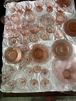 Full lot of various pink Depression glass pieces arranged on white cloth showing multiple types of cups, glasses, covered sugars, plates, and bowls.