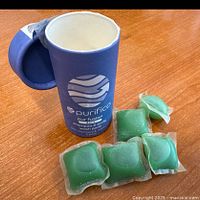 Close-up of purple-blue cylindrical container with Purifico branding and four green gel pods outside of it on wooden table.