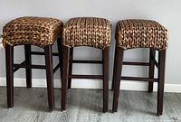 Three wood and rattan bar height stools in a row showing woven rattan seats and dark brown wooden legs on a gray floor against white wall.