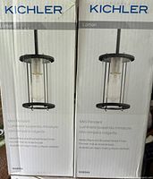 Boxes showing Kichler Loman mini pendant lights, one in matte black and the other brushed nickel, each with clear glass cylinder shade.