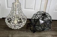 Two Pottery Barn pendant light fixtures shown side-by-side on floor, one white metal floral and one black metal geometric glass pendant