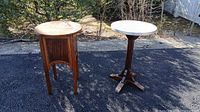 Two side tables placed outside on asphalt, one wooden cabinet-style and one round white-top pedestal table.
