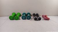 Front-facing image showing four pairs of dumbbells arranged side by side: light green Tone Fitness 3 lb, medium green Bollinger 3 lb, black NordicTrack 3 lb, and pink Cap Barbell 1 lb.