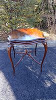 Front angled view of a dark stained wooden side table showing scalloped top and curved cabriole legs on an asphalt background.