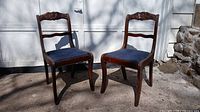 Pair of two antique wooden chairs with carved rose motifs on backrest and blue upholstered seats, placed outside on a concrete surface.