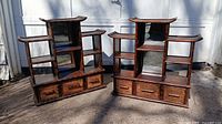 Pair of dark wood pagoda-shaped shelves with drawers and mirrors on middle shelf backs, shown outside against white wall.